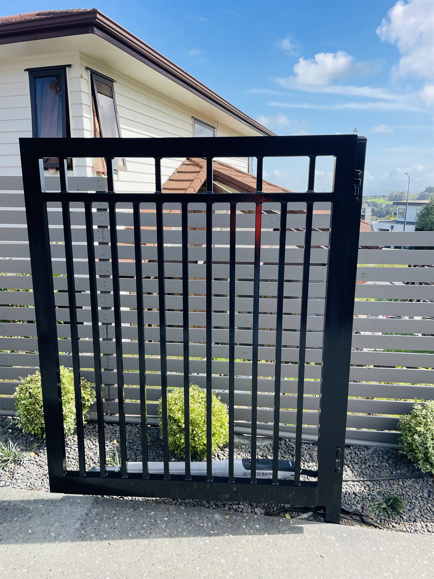 Black metal pedestrian gate against a repainted grey slat fence