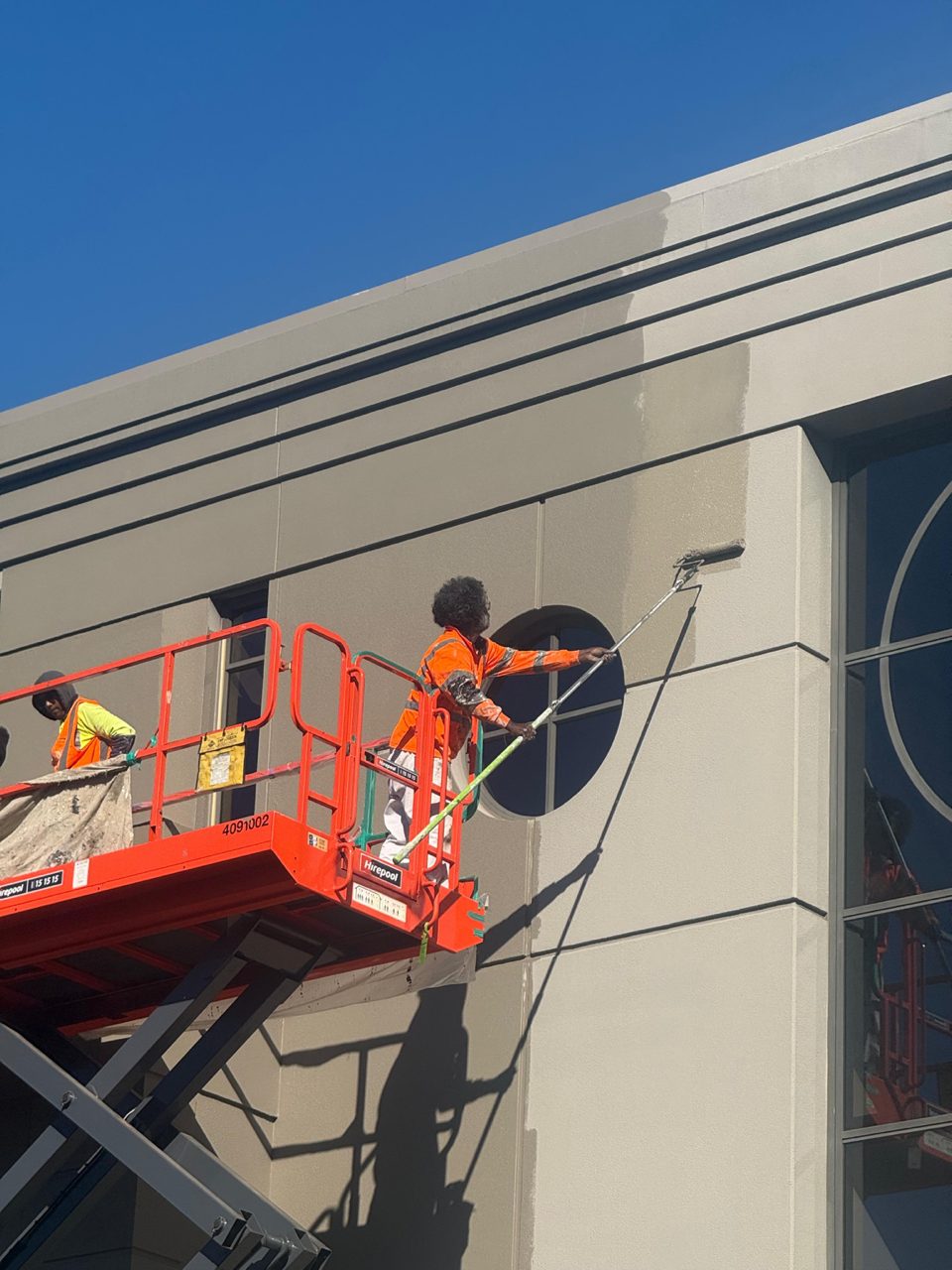 Painter on a scissor lift rolling coating onto the upper facade in high-visibility gear